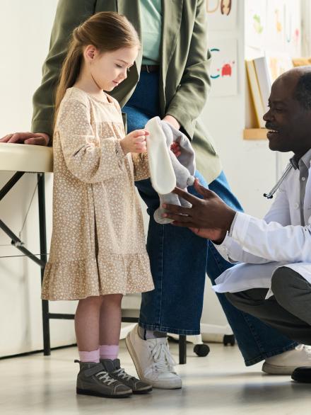 Senior doctor kneeling and assisting young girl holding white stuffed animal in medical office environment with diverse people present and medical equipment surrounding